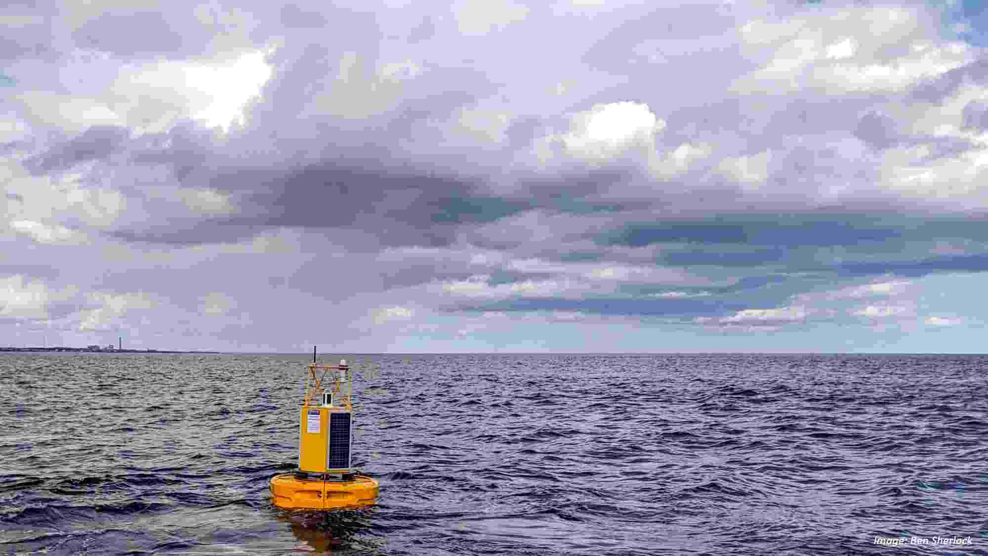 A photograph of a calm sea on a cloudy bay, with a large bright yellow marker buoy in the foreground.